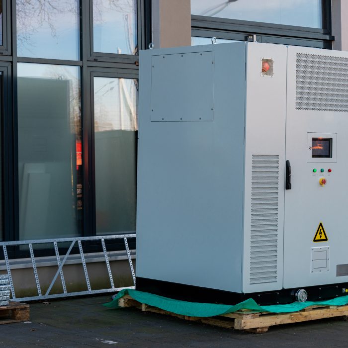 A large industrial cooling unit stands on a green mat outside a contemporary building. The unit features control panels, warning signs, and is surrounded by metal scaffolding and windows.