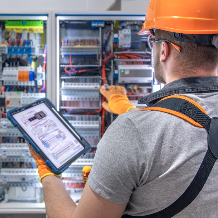 A male electrician in overalls, focused on work in switchboard with fuses, using tablet. High quality photo. An electrician with a tablet in his hand works in the switchboard. Copy space.