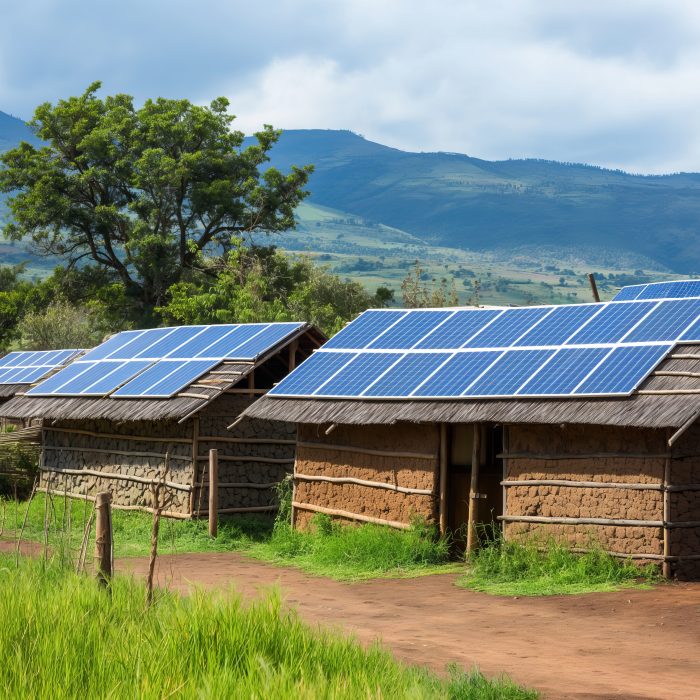 Several mud houses with thatched roofs in an African village, equipped with solar panels, against a background of trees and mountains